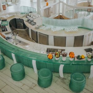 An aerial view of a modern, semi-circular bar with a green glass counter surrounded by matching stools at a Maldives luxury resort. The bar is equipped with various appliances, glassware, and a selection of fruits. The setting is bright and airy, with light wood accents and a spiral staircase.