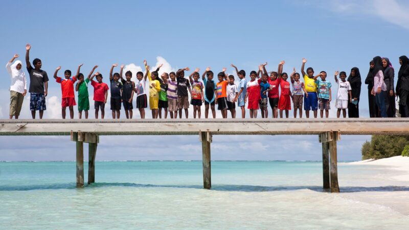 A group of children and adults stand on a wooden pier above the clear, turquoise waters of the Maldives. Everyone is wearing casual attire and waves at the camera. The sky is clear and sunny, and a sandy beach with greenery from Namoona is visible in the background.
