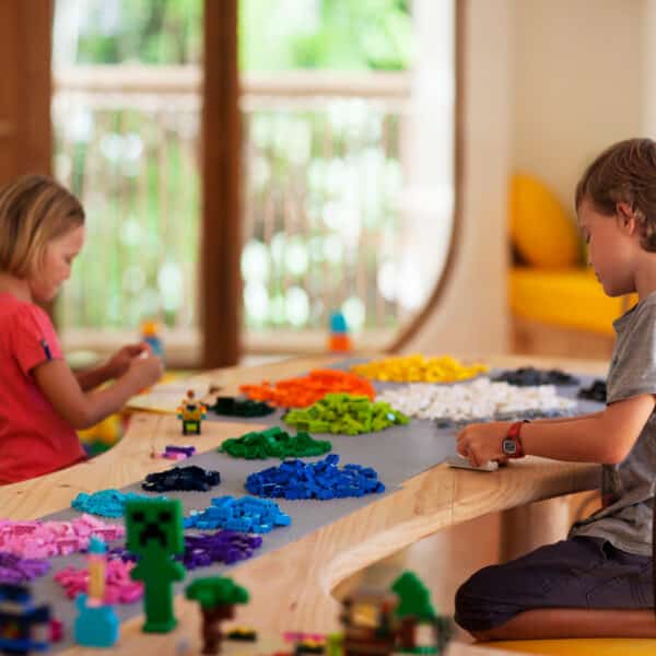 Two children, a girl and a boy, sit at a wooden table covered with assorted colorful LEGO pieces. The girl, wearing a red shirt, is focused on building with the LEGO. The boy in a grey shirt also engages with the LEGO. Large, bright windows flood the room with light, creating an atmosphere reminiscent of a Soneva Maldives exclusive experience.