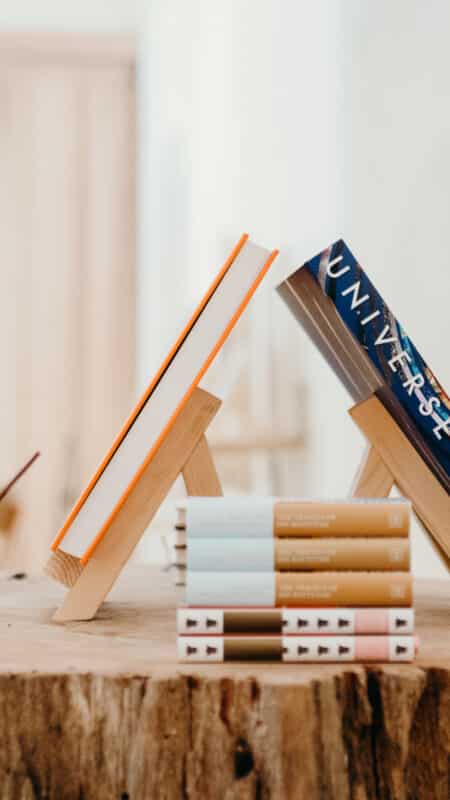 A wooden table displays various books. Two books are propped up on small wooden stands, including one titled &quot;Universe.&quot; Additional books are stacked beside and in front of the stands. Softly lit shelves with more items, reminiscent of a serene Maldives holiday destination, are visible in the blurred background.