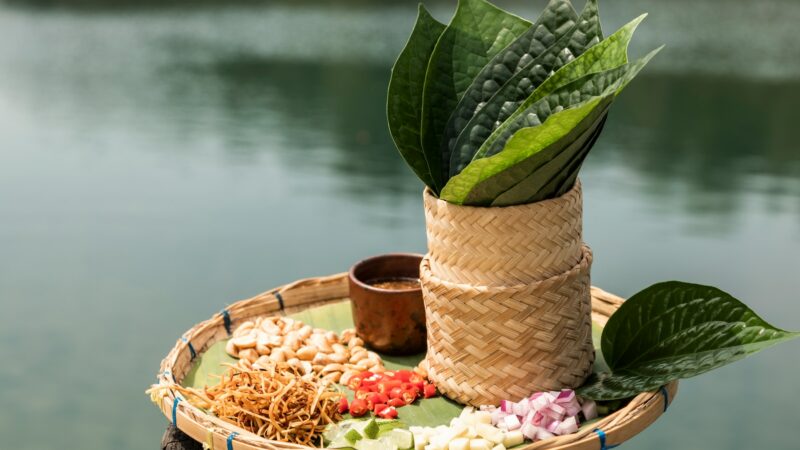 A round woven tray holds various ingredients atop a banana leaf, including sliced red chilis, garlic, shallots, roasted peanuts, and dried shrimp. Upright betel leaves rest in a small woven basket by the water. This is part of a Thailand Exclusive Experience at Soneva.