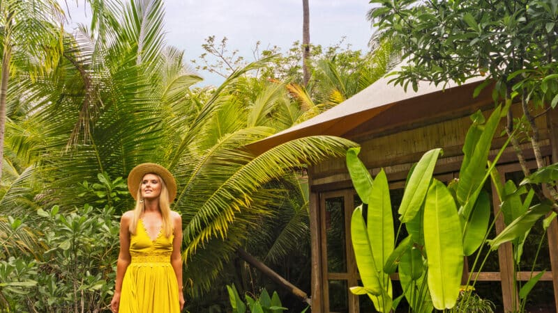 A woman in a yellow dress and hat stands on a wooden walkway surrounded by lush tropical plants and trees, with a palm tree and a wooden building in the background. Enjoy this Thailand exclusive experience under the partly cloudy sky, epitomizing the luxury of Soneva resorts.