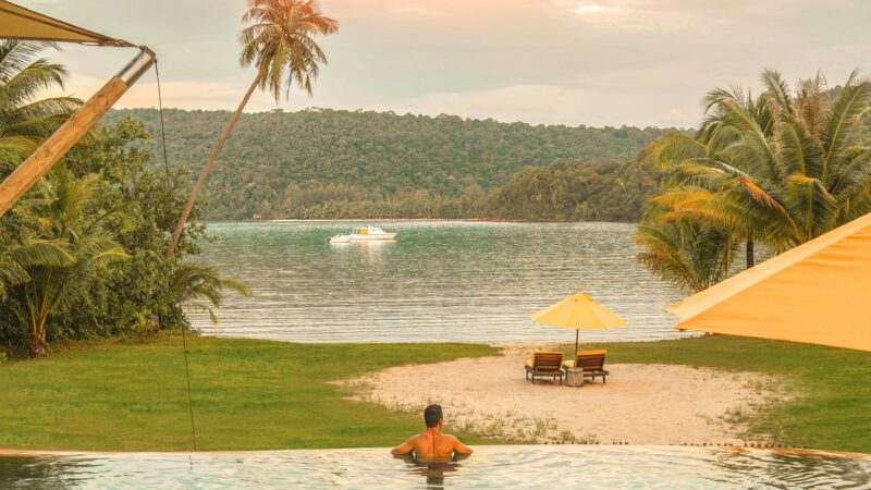 A person enjoying a serene view while standing in an infinity pool at Soneva Kiri Resort, overlooking a calm lake with a small boat in the distance. The scene is surrounded by lush greenery and palm trees, with lounge chairs and umbrellas on the grass and sandy shore. The sky is painted with warm tones of a sunset.