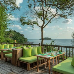 A scenic outdoor restaurant overlooks the ocean, featuring wooden tables and green cushioned seating. Lush vegetation and trees surround the deck, enhancing the tropical vibe under a partly cloudy blue sky. Take advantage of Soneva Exclusive Offers for an unforgettable dining experience.