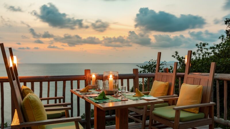 A cozy outdoor dining setting on a wooden deck overlooks the ocean at sunset, offering an exclusive experience in Thailand. The table is set with yellow cushioned chairs, green napkins, candles in glass holders, and tableware. Fluffy clouds dot the sky, with greenery visible on the right—a true Soneva escape.