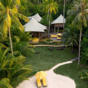 A serene beachfront resort set within lush greenery, featuring Soneva Exclusive Offers. Multiple white canvas-topped tents are surrounded by palm trees. Two yellow lounge chairs sit under a yellow parasol on a sandy pathway leading to the entrance, offering a tranquil and tropical atmosphere.