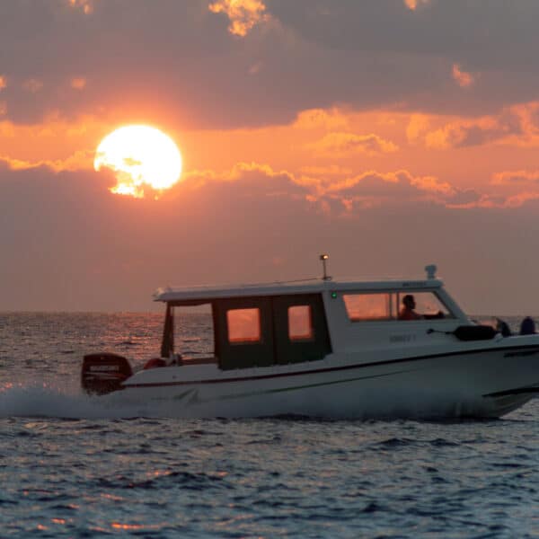 A boat speeds across the ocean at sunset, with the setting sun partially hidden by clouds. The sky is painted in shades of orange and pink, casting a warm glow on the waves—truly a Maldives exclusive experience that captures the essence of Soneva&#039;s unparalleled beauty.