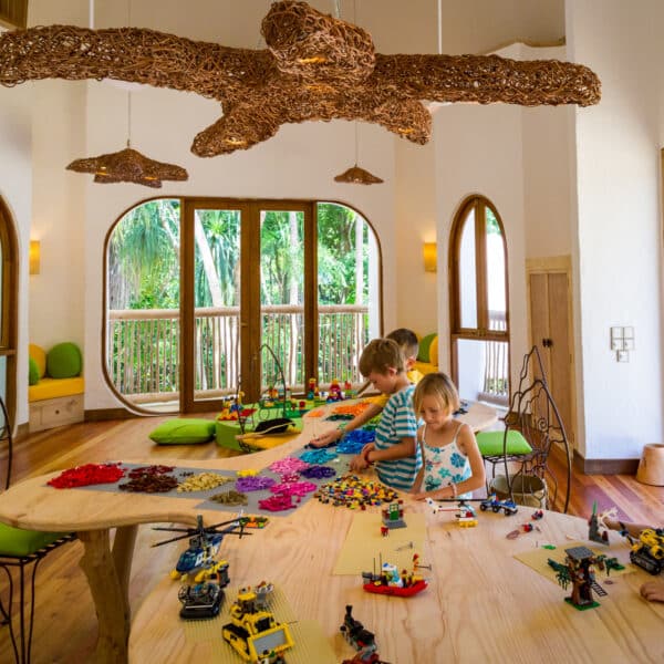 Children are sitting around a large wooden table in a brightly lit room with large arched windows, reminiscent of a Maldives exclusive experience. The whimsical chandelier illuminates their playful scene as they engage with colorful building blocks and small toy vehicles, fostering a creative atmosphere.