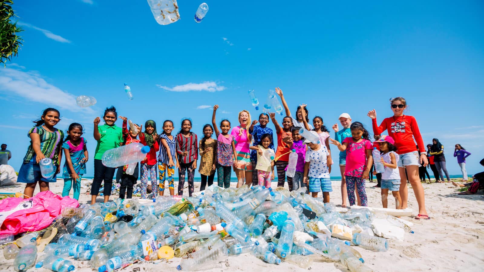 A group of people, including children and adults, stand on a beach in the Maldives Namoona behind a large pile of collected plastic bottles. They are cheerfully raising their hands and some are throwing bottles into the air. The sky is clear with a few clouds contrasting the blue sky.