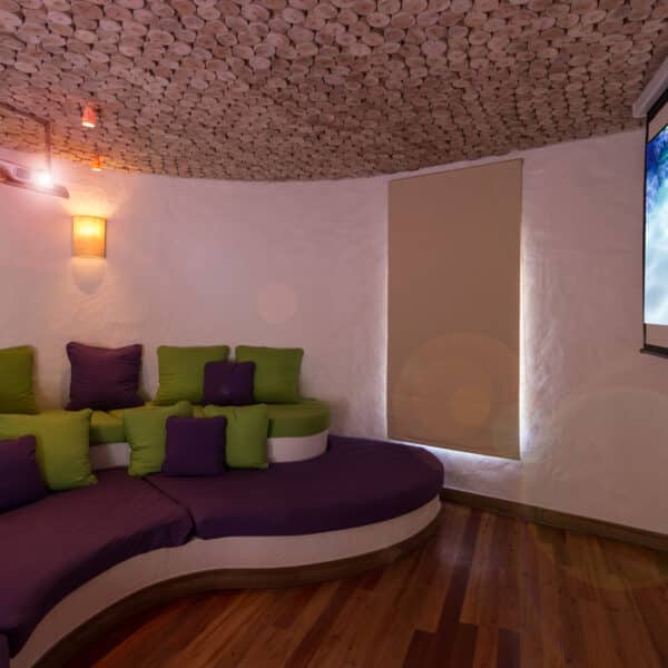 A cozy home theater with a curved seating area filled with purple and green cushions. The room has warm lighting, a wooden ceiling with circular patterns, and a large screen displaying an animated fish character. This Maldives Exclusive Experience is complemented by a window covered by a beige blind.