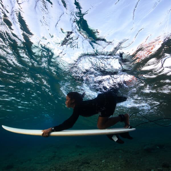 Underwater photograph of a person wearing a black wetsuit, duck diving with a white surfboard through a wave. The surface of the water captures reflections and sunlight, while the seabed below is visible. This Maldives exclusive experience at Soneva offers breathtaking views above and below the waterline.