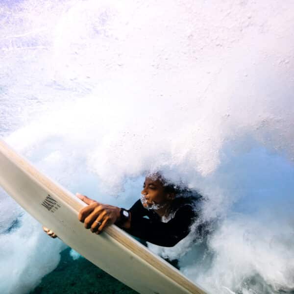 A surfer is submerged underwater, holding onto a surfboard while surrounded by swirling bubbles and whitewater. The image captures the dynamic motion and powerful force of the ocean. The surfer, wearing a black wetsuit, appears calm and composed, enjoying their Maldives Exclusive Experience at Soneva.
