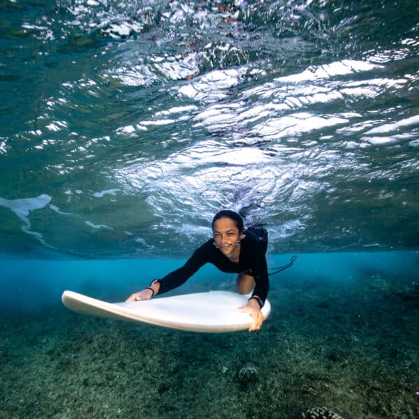 A person underwater smiles while holding a surfboard. Dressed in a dark swimsuit and close to the ocean floor, which is visible through the clear blue water above them, they enjoy an exclusive experience reminiscent of the Maldives. The surface of the water ripples, capturing sunlight from above.