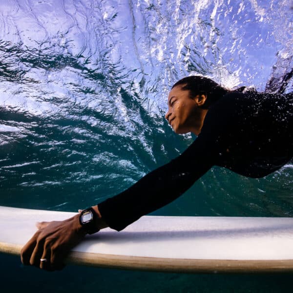 Underwater view of a person surfing in the Maldives, wearing a black wetsuit and a watch on their left wrist. The person is holding onto the edge of the surfboard, with bubbles and water currents visible above. This Soneva exclusive experience captures the dynamic and immersive nature of underwater surfing.
