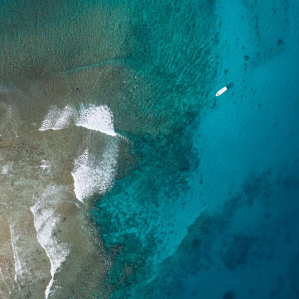 Aerial view of a pristine coastline with clear turquoise water gradually transitioning to deeper blue. The sandy shore has patches of white, appearing as waves, and a small white boat floats near the darker waters. This serene scene is reminiscent of a Maldives exclusive experience at Soneva.