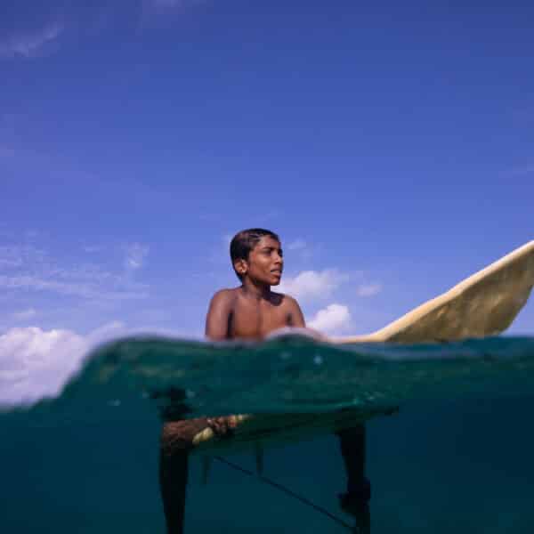 A boy, with dark hair and skin, sits on a surfboard in the ocean under a clear blue sky. The photo is split between above and below the waterline, showing the water's surface and the underwater part of the surfboard. He looks forward with a calm expression, embodying a Maldives exclusive experience at Soneva.