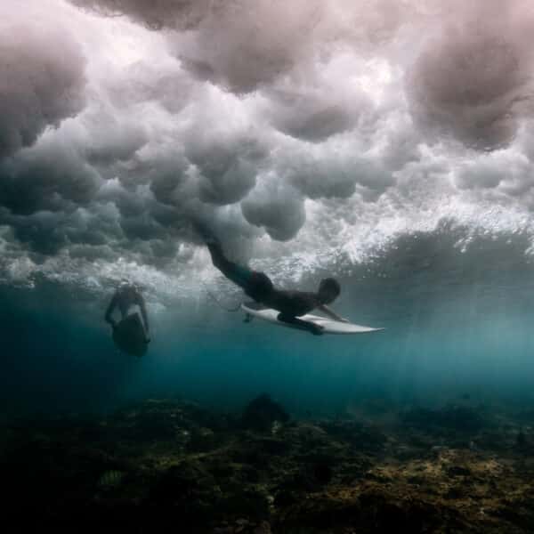 Underwater view of two surfers in the ocean just below the water's surface. One surfer is in motion with a surfboard, while the other appears to be diving or swimming. Above them, turbulent waves create dramatic, frothy patterns. Enjoy this Maldives exclusive experience at Soneva, where the seafloor is dimly lit.