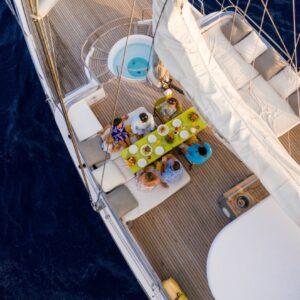 Aerial view of a group of people dining on a sailboat during their Maldives luxury yacht holiday. The boat's wooden deck is adorned with a yellow tablecloth under a sail. Plates and drinks are set on the table, surrounded by cushioned seating. The boat is in deep blue water.