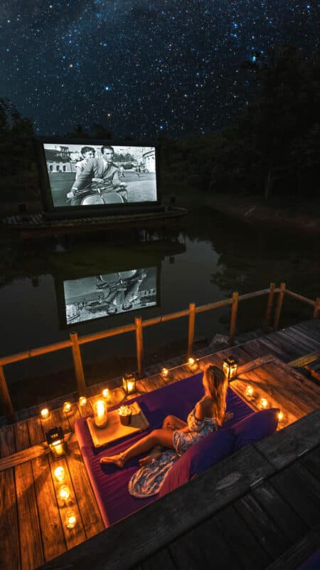 A woman in a summer dress sits on a dock surrounded by lanterns, watching a black-and-white movie on a screen across a small lake. The night sky above is filled with stars, reflecting on the calm water, creating a serene and picturesque outdoor cinema scene—an exclusive Thailand experience by Soneva.