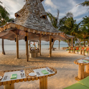 A tropical beach setting with a thatched-roof gazebo, surrounded by palm trees. Wooden tables set with plates and cutlery are arranged nearby on sandy ground. Cushioned seating along a curved bench with a scenic ocean backdrop completes the serene ambiance, epitomizing Soneva Maldives luxury.