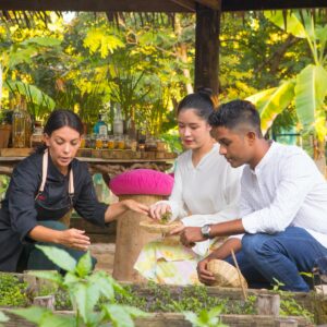 Three people are gathered around a garden bed under a wooden pavilion. One woman, dressed in black, points to the plants, while the other two, a man and a woman in white shirts, listen attentively and hold small wicker baskets—a true Thailand exclusive experience at Soneva. In the background, there is a table with plants.