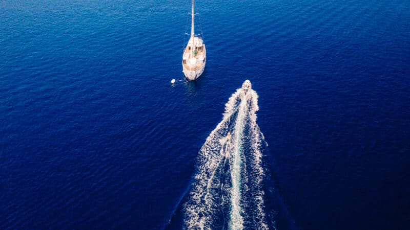 Aerial view of a sailing boat anchored in the deep blue sea during a Maldives luxury yacht holiday at Soneva, with a smaller motorboat speeding away, leaving a trail of white waves behind. The calm, expansive ocean contrasts with the dynamic motion of the motorboat.