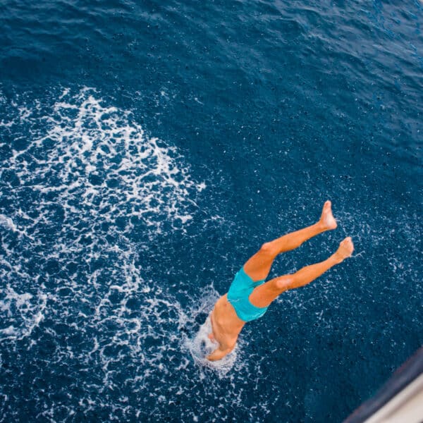 A person wearing turquoise swim trunks is diving headfirst into the blue ocean from the side of a luxury yacht, creating splashes in the water during their Maldives holiday at Soneva. Only the lower half of their body is visible, with legs pointed upwards.