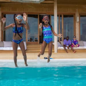 Two young girls wearing blue swimsuits and goggles are excitedly jumping into a pool, creating splashes. They are on a wooden deck of a modern building with large glass doors, reminiscent of a Maldives Exclusive Experience. Three people relax on a bench in the background, watching the girls.