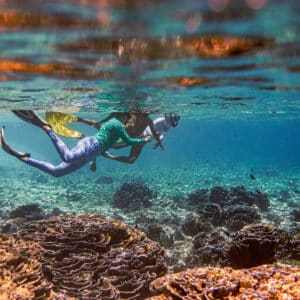 A person wearing snorkeling gear swims underwater near a vibrant coral reef during a family holiday at Maldives' Soneva resort. The clear water reveals the colorful coral formations below, while sunlight from above creates a serene and visually striking scene.