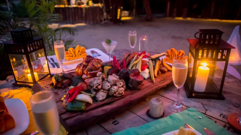 A festive outdoor table setting with a variety of grilled meats, vegetables, and red peppers arranged on a wooden platter. The scene is illuminated by lanterns and candlelight, with champagne glasses placed around the table, creating a warm and inviting atmosphere—truly a Thailand Exclusive Experience at Soneva.