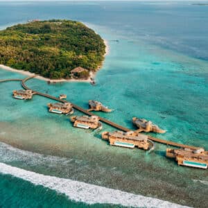 Aerial view of an island resort surrounded by blue waters. The lush island, connected by wooden walkways to overwater bungalows extending into the sea, looks like paradise. A sailboat is visible in the distance as waves break near the shore. Discover Soneva Exclusive Offers for a luxurious getaway.