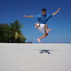 A person wearing a blue shirt and white shorts jumps and performs a skillful kick with a soccer ball on a sandy beach during their family holiday at Soneva in the Maldives. Palm trees, beach chairs, and clear blue sky are in the background, while the person&#039;s shadow is visible on the sand.