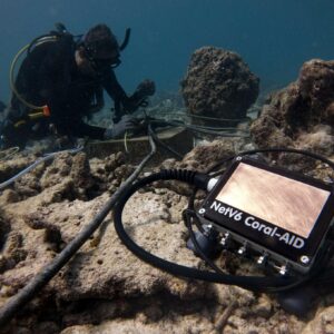A scuba diver works underwater near a rocky reef, operating a device labeled "Nev6 Coral-AID." Connected by cables, the device seems to be part of a coral restoration project by Maldives Foundation in collaboration with Soneva. The surrounding area consists of vibrant coral and submerged rocks.