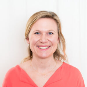 A woman with blonde hair is smiling at the camera. She is wearing a coral-colored top and a delicate necklace. The background is a plain white wall.