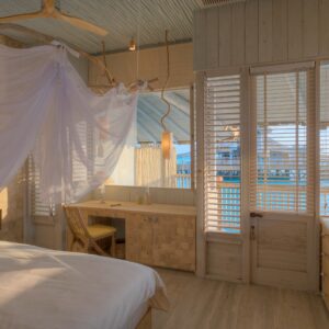 A serene bedroom with a wooden interior features a canopy bed draped in white fabric. A large window with shutters offers a view of an overwater bungalow and the ocean, reminiscent of Maldives luxury. The room includes a desk and chair, shelves with neatly placed items, and warm natural lighting.