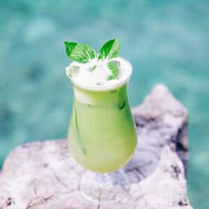 A refreshing green drink garnished with mint leaves in a tall glass, placed on a natural wooden surface with a clear turquoise water backdrop. The drink looks inviting and summery against the vibrant blue-green background, reminiscent of Soneva Exclusive Offers.