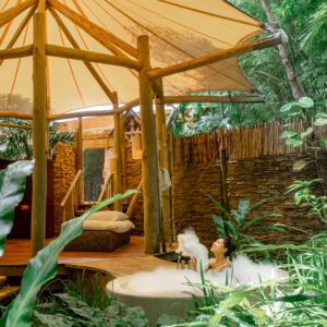 A woman relaxes in an outdoor bathtub filled with bubbles, surrounded by lush green foliage. The area features wooden structures with a tent-like canopy and stone walls, blending seamlessly with the natural environment. She appears tranquil and immersed in the serene setting, enjoying Soneva Exclusive Offers.