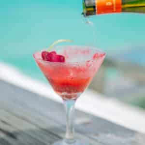 A refreshing red frozen drink garnished with a cherry and lemon twist, served in a frosty martini glass on a weathered wooden surface. An orange-capped bottle is being poured into the glass. In the background, a blurred turquoise sea shimmers under bright sunlight, hinting at Soneva Exclusive Offers.