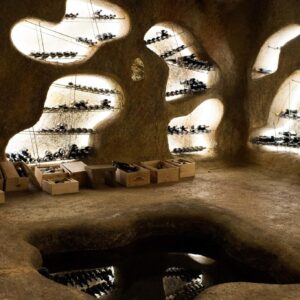 A rustic wine cellar with organic, cave-like walls featuring recessed, backlit shelves filled with wine bottles. Wooden crates of wine are scattered on the earthy floor, evoking an ancient and natural ambiance reminiscent of the luxurious simplicity found at a Maldives resort like Soneva.