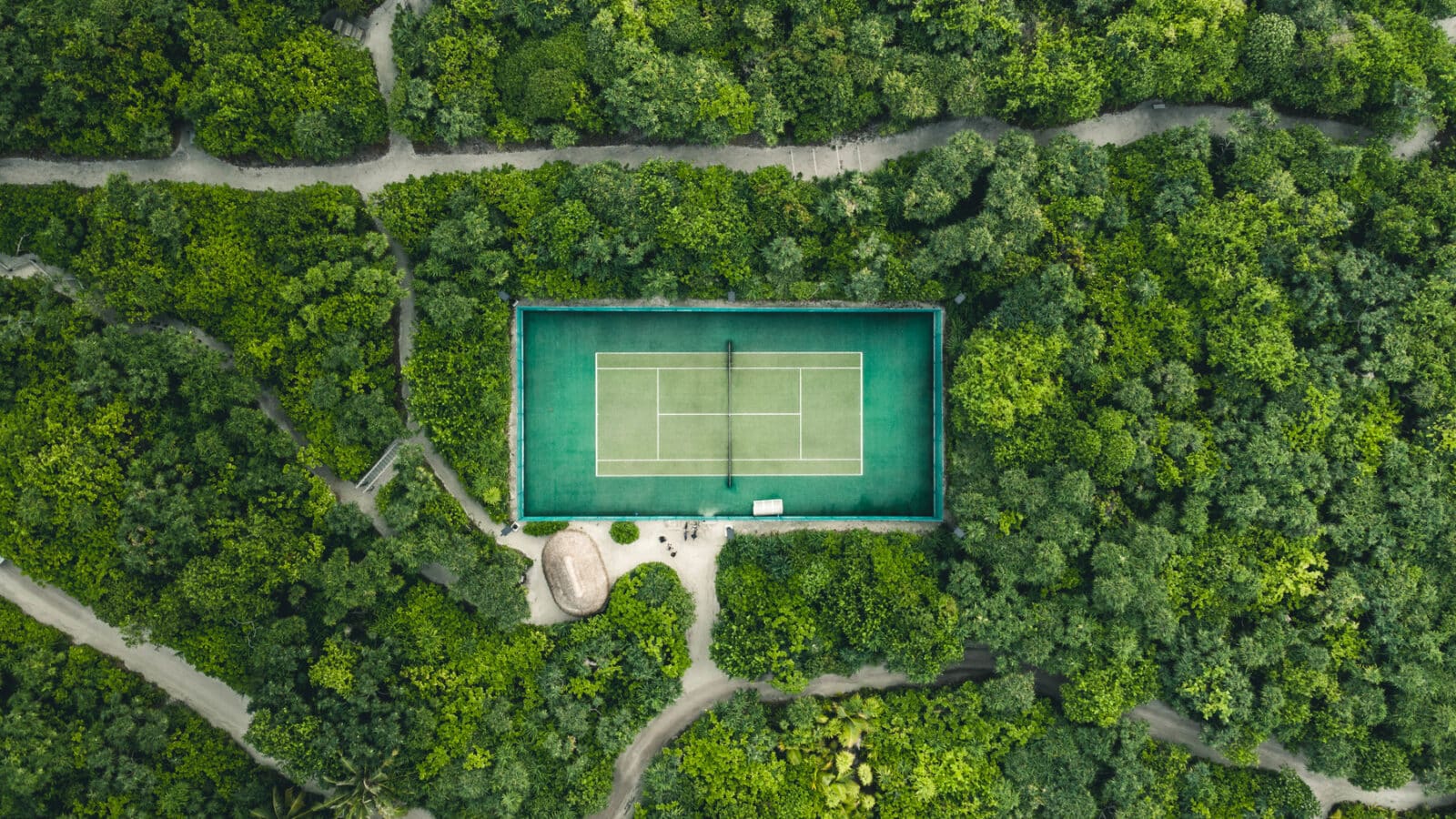 Aerial view of a tennis court surrounded by dense, lush green forest. Pathways meander through the trees, leading to the court, which stands out with its vibrant green surface. A small hut is visible near the court&#039;s lower left corner, reminiscent of a Maldives exclusive experience at Soneva resorts.