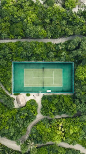 Aerial view of a tennis court surrounded by dense, lush green forest. Pathways meander through the trees, leading to the court, which stands out with its vibrant green surface. A small hut is visible near the court&#039;s lower left corner, reminiscent of a Maldives exclusive experience at Soneva resorts.
