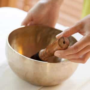 Close-up of a person holding a Tibetan singing bowl, with both hands on either side of the bowl and a wooden mallet resting inside. The setting appears calm and serene, suggesting a meditative or healing atmosphere, reminiscent of Soneva Exclusive Offers' tranquil retreats.