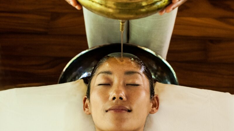 一个人闭着眼睛躺着，正在接受阿育吠陀精油护理，这是 Soneva 独家优惠的一部分。一股精油正从他们额头上方的黄铜容器中倾泻而出。背景是木地板，人身上盖着白色毛巾。