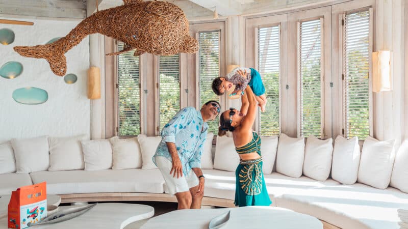 A family of three enjoys a moment in a sunlit room with large windows, reminiscent of a tranquil Maldive Soneva retreat. The father, in a blue shirt and white shorts, smiles as the mother, wearing a green dress, playfully lifts their young child. A decorative woven dolphin hangs from the ceiling.