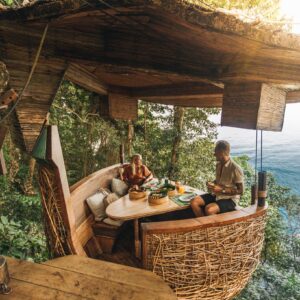 Two people are seated in a cozy, elevated treehouse overlooking the ocean. The treehouse has a rustic wooden design with wicker accents and a seating area adorned with cushions. The surroundings are lush, green forest with a view stretching out to the blue water—truly embodying Soneva Exclusive Offers.