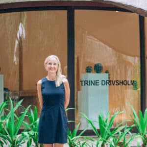 A person with long blonde hair, wearing a sleeveless black dress, is standing outdoors in front of a building. Behind them, a glass window displays "TRINE DRIVSHOLM." Lush green plants are in the foreground, creating a scene that evokes the exclusivity of a Maldives experience like Soneva.