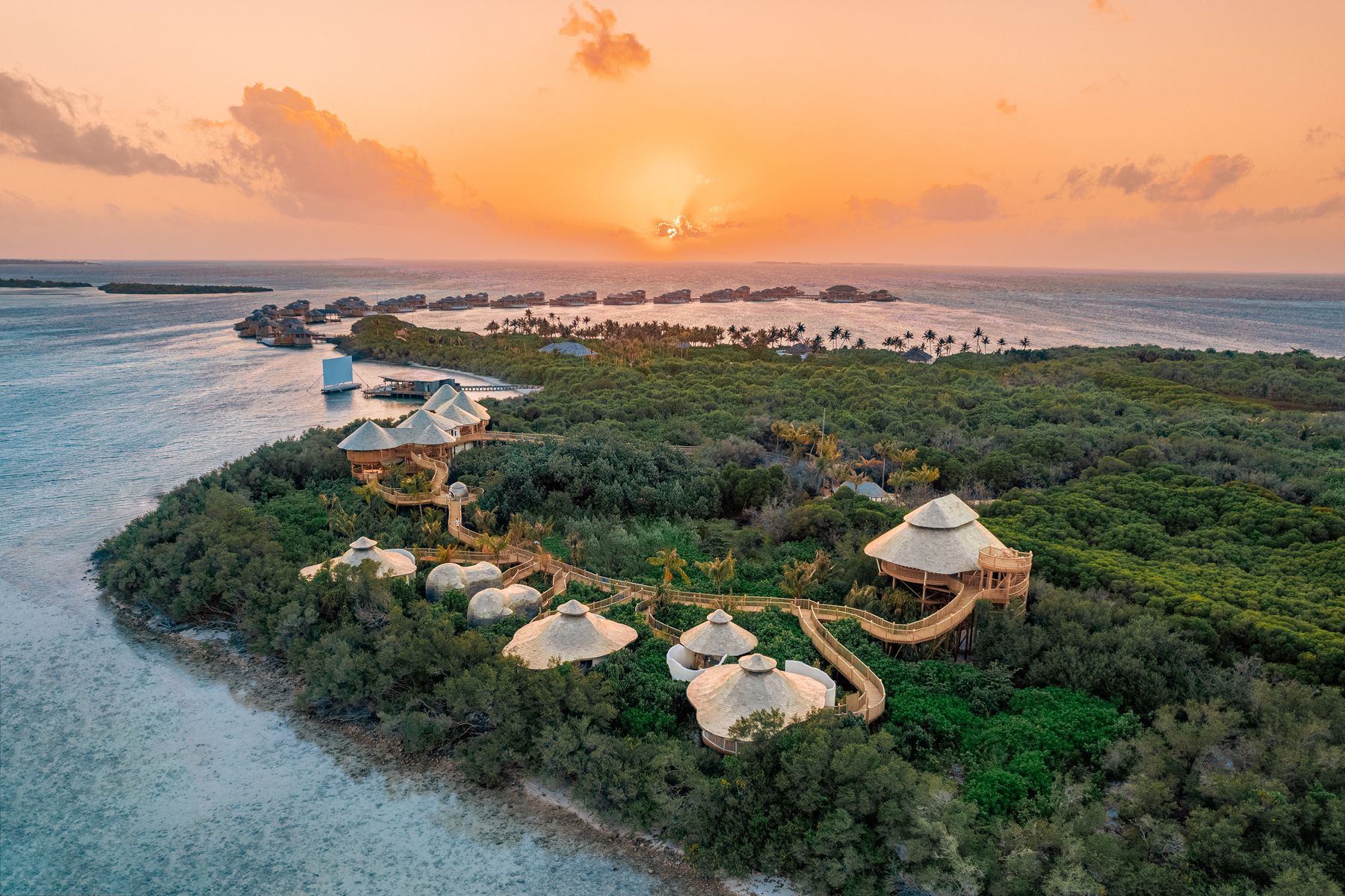 An aerial view of a tropical resort island at sunset reveals the Maldive Spa Wellness experience, with lush greenery, interconnected wooden huts with thatched roofs, and clear blue waters. Overwater bungalows and small islands dot the horizon under a colorful sky.