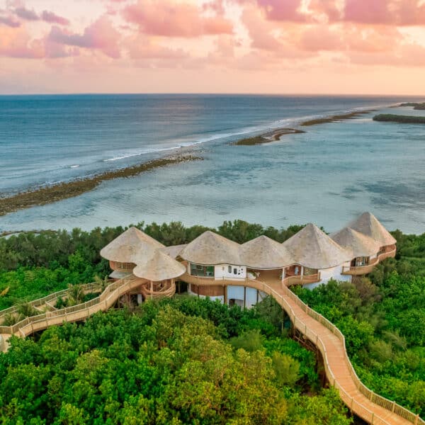 Aerial view of a tropical seaside resort featuring round, thatched-roof bungalows connected by wooden walkways, surrounded by lush green vegetation. The resort, including a renowned Maldive spa, overlooks a picturesque coastline with calm turquoise waters and a colorful sunset sky.