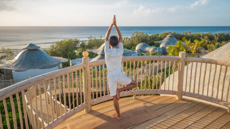 A person practices yoga in the tree pose on a wooden deck overlooking the ocean. The deck, part of Soneva Exclusive Offers, features a wooden railing and has greenery and dome-shaped structures in the background. The sky is clear with a few clouds.
