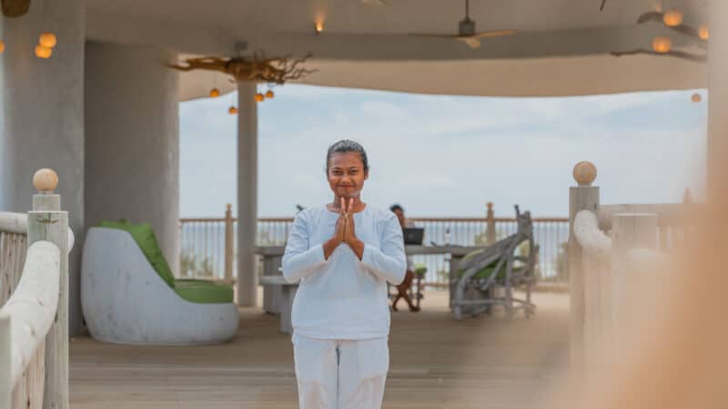 A person in a white outfit stands with hands together in a prayer-like gesture, smiling gently. The background features an outdoor, covered wooden structure with seating and decor shaped like tree branches, overlooking a scenic view reminiscent of a Maldives spa wellness retreat.